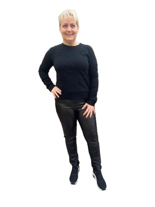 Person wearing a black long-sleeve top and leather pants standing in front of a brick wall.