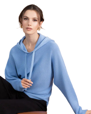 Woman wearing a blue hoodie sitting on a brown leather couch with a wooden paneled wall in the background.