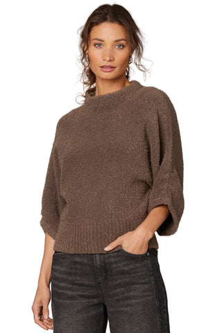 Woman wearing a brown sweater and dark jeans against a neutral background