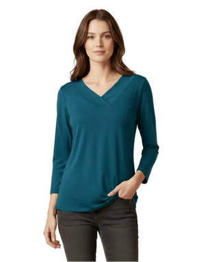 A woman modeling a teal long-sleeve V-neck top with dark gray pants, standing against a solid beige studio background.
