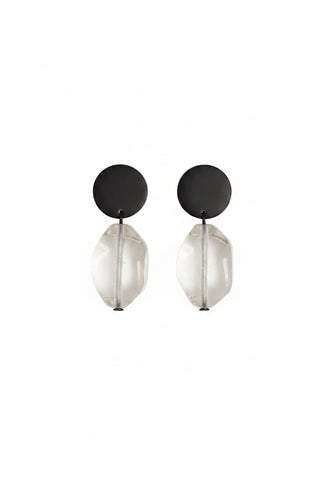 A pair of minimalist drop earrings featuring matte black circular studs and faceted clear glass teardrop pendants, displayed on a plain off-white surface.