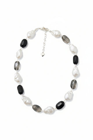 A beaded necklace with irregular white baroque pearls, faceted black gemstone beads, and clear glass beads on a silver chain with a small heart charm, displayed on a mottled brown granite surface.