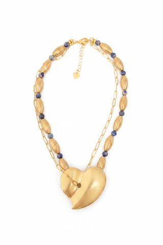 A gold-toned beaded necklace with oval wooden beads and small dark blue beads, featuring a large sculptural gold pendant, displayed on a textured gray stone surface.