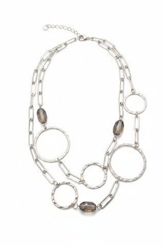 A silver-toned geometric chain necklace with large open-link chains, circular disc charms, and small clear faceted beads, displayed on a mottled brown countertop.