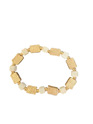 A beaded stretch bracelet featuring textured gold-toned rectangular beads, small round gold spacer beads, and off-white square beads, displayed on a mottled brown and gray stone surface.