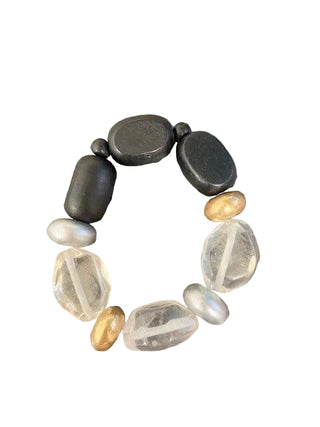 A handmade beaded bracelet featuring smooth black stones, faceted clear quartz beads, and small metallic gold and silver beads, displayed on a textured brown stone surface.