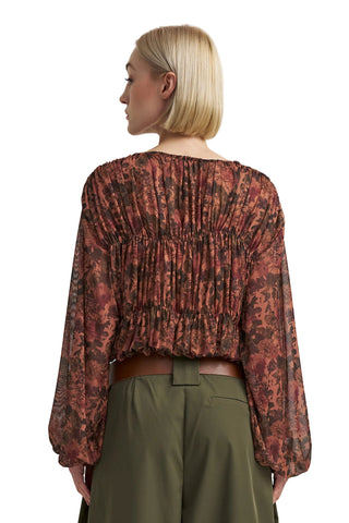 A woman shown from the back wearing a long-sleeve, gathered blouse with an abstract floral pattern in rust red, brown, and black, paired with olive green trousers against a light gray studio background.
