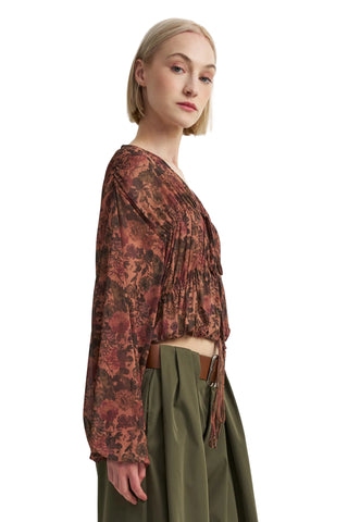 A woman shown in side profile wearing a long-sleeve, gathered wrap-style blouse with an abstract floral print in rust, brown, and burgundy tones, paired with olive green high-waisted trousers.