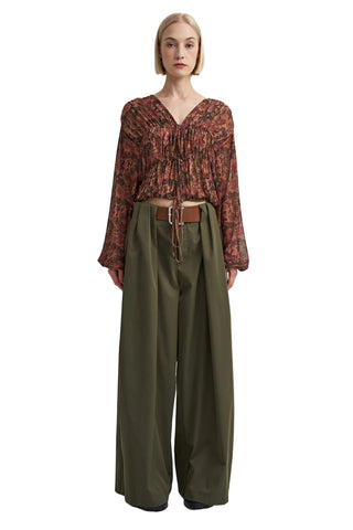 A model stands against a plain light gray studio background wearing a long-sleeve, gathered V-neck blouse with an abstract rust and brown pattern, paired with wide-leg olive green trousers and a brown belt.