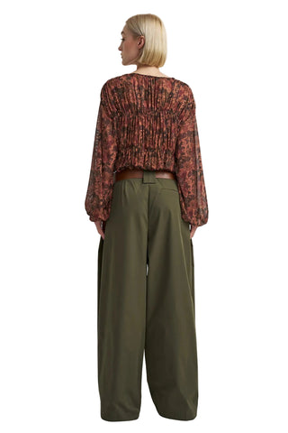 A model shown from the back wearing a long-sleeve, gathered, cropped blouse with a rust-red and black floral pattern, paired with olive green wide-leg trousers, against a plain light gray background.