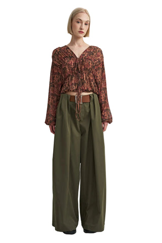 A model stands against a plain light gray studio background wearing a rust-brown patterned long-sleeve top with a tie neckline and wide-leg olive green trousers with a brown belt.