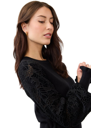 A woman wearing a black long-sleeve top with intricate floral lace applique on the sleeves, shown against a plain light gray studio background.