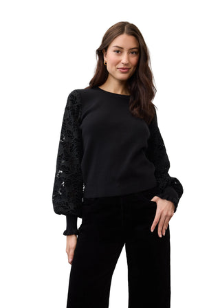 A woman modeling a black long-sleeve top with textured lace sleeves, paired with black pants, against a plain light gray studio background.