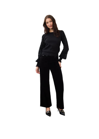 A woman modeling a black long-sleeve knit top with textured sleeves and black wide-leg trousers, standing against a plain white wall on a concrete floor.