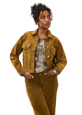A model wearing a mustard yellow corduroy jacket and matching corduroy pants over a floral print top, standing against a plain off-white wall.