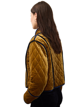 A woman shown from the side and back wearing a mustard yellow quilted velvet jacket with dark navy piping, set against a plain off-white background.