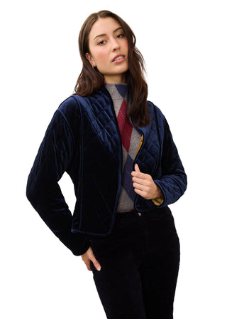 A woman modeling a navy blue velvet cropped quilted jacket over a gray and red argyle sweater, paired with black pants, against a plain off-white studio background.