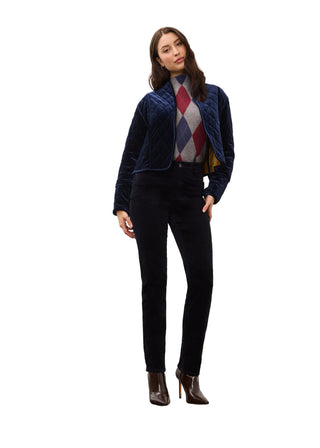 A woman models a navy blue quilted zip-up jacket over a multicolor argyle sweater, paired with dark trousers and brown ankle boots, standing against a plain off-white wall.
