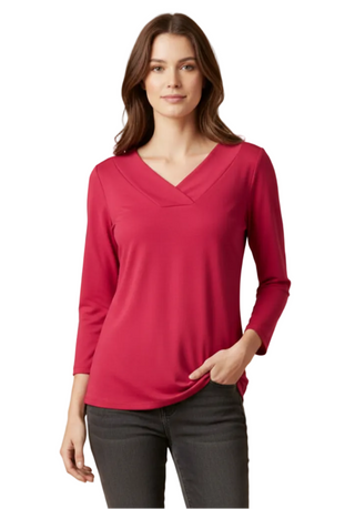 A woman modeling a long-sleeve V-neck top in a rich magenta pink color, paired with dark gray jeans, against a solid beige studio background.