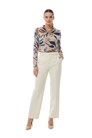 A model stands against a plain white studio background wearing a long-sleeve cowl-neck top with a beige, blue, and brown botanical pattern, paired with off-white wide-leg trousers and nude heels.