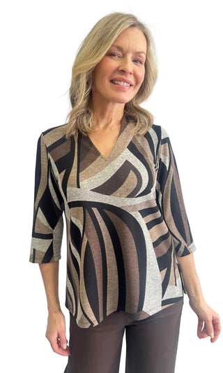 A woman modeling a three-quarter sleeve wrap-style top with a geometric abstract pattern in black, brown, beige, and gray, paired with dark brown pants, against a light gray studio background.