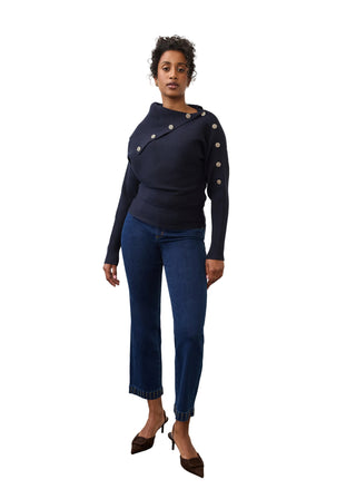 A model stands in a studio wearing a dark navy long-sleeve knit top with asymmetrical gold-tone buttons and dark blue cropped jeans with contrast stitching, paired with dark brown pointed-toe heels.