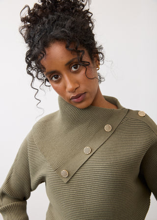 A close-up shot of a model wearing an olive green ribbed knit turtleneck sweater with asymmetrical decorative buttons, set against a plain light background.