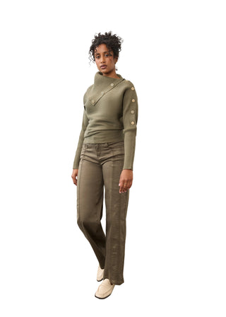 A model stands in a studio wearing an olive green asymmetrical long-sleeve knit top with gold-tone buttons down one sleeve, matching olive green pants, and cream-colored loafers.