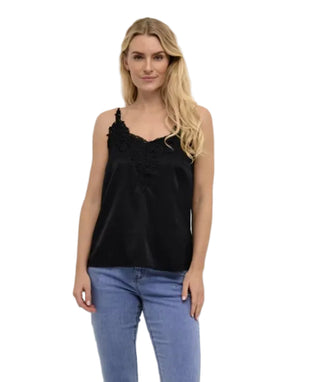 A blonde woman wearing a black satin camisole top with floral lace applique on the neckline, paired with light blue jeans, standing against a plain white wall.