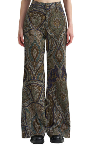 A model is shown from the waist down wearing high-waisted, wide-leg trousers with an intricate paisley pattern in olive green, brown, gold, and navy blue, paired with black shoes against a plain white studio background.