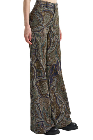 A model is shown from the waist down wearing high-waisted, wide-leg trousers with an intricate paisley pattern in olive green, brown, gold, and purple. The model is also wearing black leather shoes against a clean white studio background.