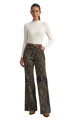 A woman modeling a white ribbed mock-neck long-sleeve top and high-waisted, wide-leg trousers featuring an intricate paisley pattern in brown, beige, and blue tones, styled with a thin brown belt and brown suede shoes against a plain white studio background.