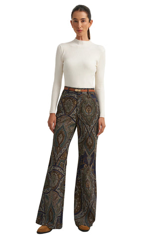 A woman modeling a white ribbed mock-neck long-sleeve top and high-waisted, wide-leg trousers with an intricate paisley pattern in brown, gold, and blue tones, paired with a thin brown belt and brown suede shoes.