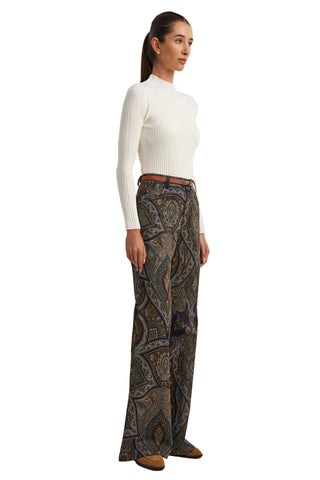 A woman stands in profile wearing a fitted white ribbed long-sleeve top and high-waisted wide-leg trousers with an intricate paisley pattern in brown, beige, and muted blue tones. She is wearing a thin brown belt and brown shoes against a plain light gray studio background.