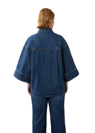 A model viewed from the back wearing a dark blue denim jacket with wide, draped sleeves and matching wide-leg denim pants, set against a plain off-white wall.