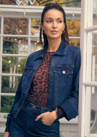 Woman wearing a denim jacket and leopard print top standing in front of a window.