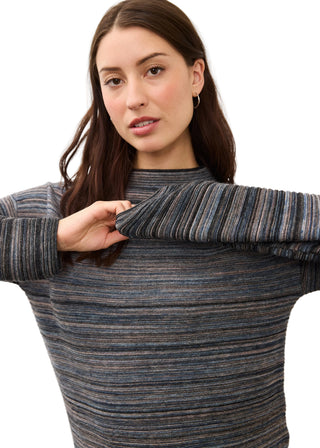 A woman wearing a ribbed knit mock neck sweater with horizontal stripes in blue, gray, and brown tones, holding up the sleeve to show the texture.