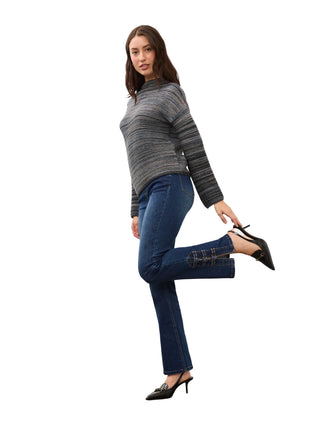 A woman models a gray and black striped mock-neck knit sweater paired with dark blue cropped jeans featuring decorative cutout details on the outer thighs, and black pointed-toe mule heels, against a light gray studio background.