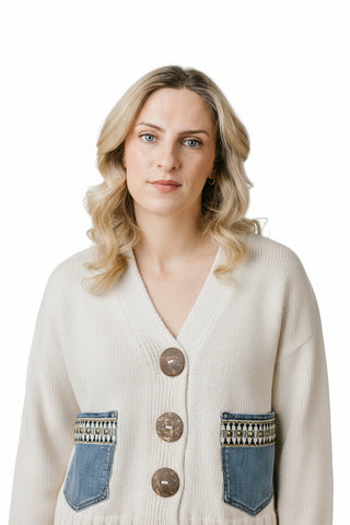 A woman wearing a cream-colored knit cardigan with blue denim patch pockets and brown buttons, standing in front of a brick wall.
