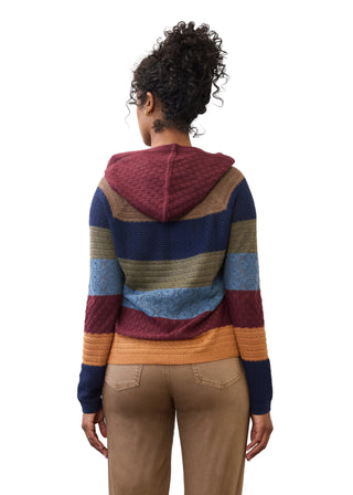 A woman viewed from behind wearing a multicolor striped knit hoodie with a maroon hood, navy, tan, blue, and mustard bands, paired with tan pants, against a plain white background.