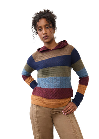 A woman modeling a multi-colored striped knit hoodie sweater with horizontal bands of navy, brown, olive green, light blue, burgundy, and mustard yellow, paired with tan pants against a plain white studio background.