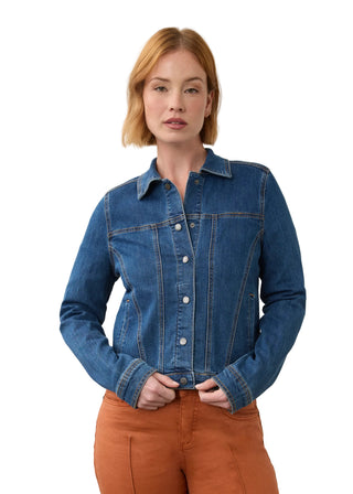 A woman with short blonde hair wearing a blue denim button-up shirt and rust-orange pants, standing against a plain light gray studio background.