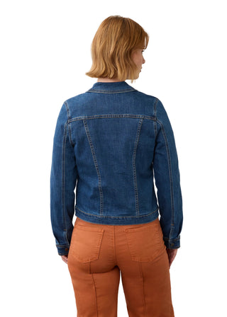 A woman viewed from behind wearing a dark blue denim jacket and rust-orange pants, standing against a plain off-white wall.