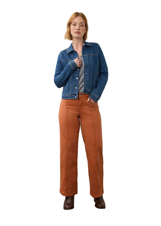A woman stands in a studio wearing a cropped blue denim jacket, rust-orange wide-leg pants, and dark brown boots against a plain off-white wall and concrete floor.