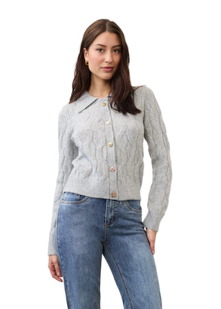 A woman modeling a light gray cable-knit cardigan with a pointed collar and decorative buttons, paired with blue jeans, against a plain light gray studio background.