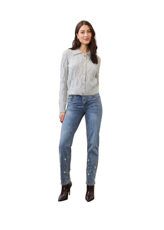 A woman stands in a studio wearing a light gray textured knit cardigan with buttons, blue jeans with small white paint splatter details near the lower legs, and dark brown heeled ankle boots.