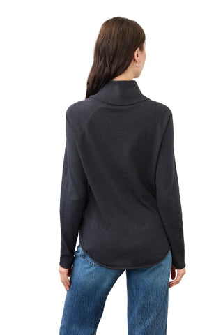 A woman viewed from the back wearing a dark charcoal gray mock neck long-sleeve top and blue jeans, standing against a plain white wall.