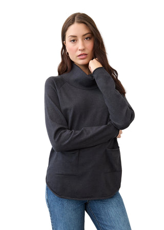 A woman modeling a dark charcoal gray long-sleeve turtleneck sweater with front patch pockets, paired with blue jeans, against a plain white studio background.