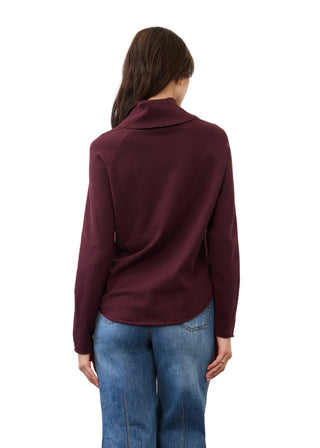 A woman viewed from the back wearing a long-sleeve mock neck top in deep burgundy and blue jeans, set against a plain light gray studio background.