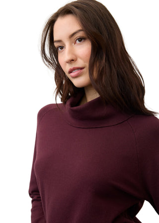 A woman with long dark brown hair wearing a solid burgundy turtleneck sweater, photographed against a clean, light gray studio background.